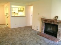 Image: The living room of an apartment. There is a small brick fireplace against one wall, and a counter cut-out window showing part of the kitchen.